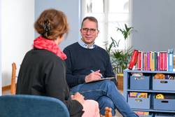 Ein Mann berät eine Frau in einem Raum der Evangelischen Beratungsstelle für Erziehungs-, Ehe- und Lebensfragen in Mülheim an der Ruhr. Der Mann sitzt mit dem Gesicht zur Kamera der Frau gegenüber, die mit dem Rücken zur Kamera sitzt. Er hat kurzes braun-graues Haar und eine Brille. Er trägt ein hellblaues Hemd unter einem dunkelblauen Pullover. Er hat ein Klemmbrett in der Hand und redet gerade. Die Frau hat rotes Haar und trägt einen schwarzen Blazer und eine rotes Tuch.
In dem Raum sind rechts Bücher in einem Regal zu erkennen, sowie diverse Utensilien für Beratungen - zum Beispiel Holzfiguren, ein Plüschtier und Spielzeug.
Im Hintergrund erkennt man eine Pflanze und eine Sitzgruppe mit Kinderstühlen.